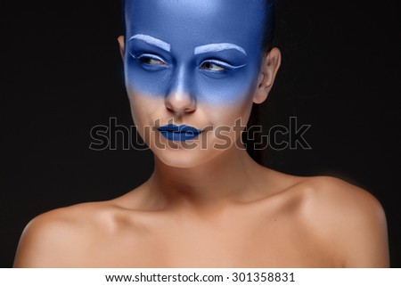 Portrait of a young woman who is posing covered with blue paint in the studio on a black background. Girl has blue lips and white eyebrows