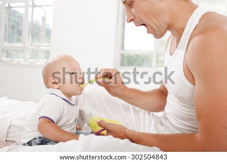 Young smiling father with his nine months old son on the bed at home on white home background. Dad feeding his son with a spoon