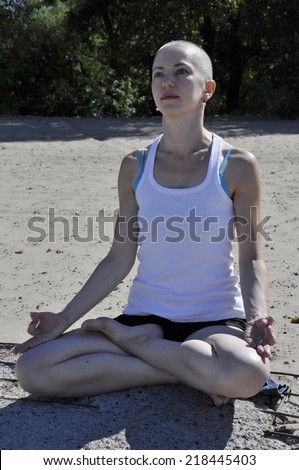 Bald woman sitting in lotus position on a beach, head against dark trees.