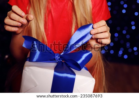 little girl whith long blond hair in red dress holding a big white gift-box with blue bow on the backgroud of holiday shining lights