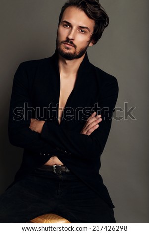 Fashion studio photo of sexy brunette handsome man dressed in a black jacket and jean whith belt sitting on the cheir,bussines
