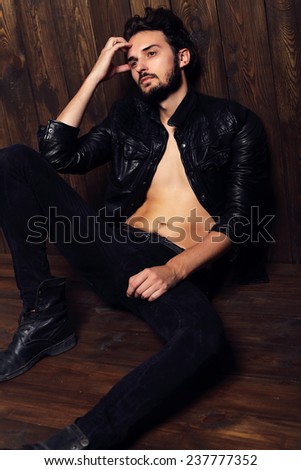 Fashion photo of sexy stylish and  handsome brunette man dressed in a black leather jacket jeans and shoes sitting on the wood flor and posing