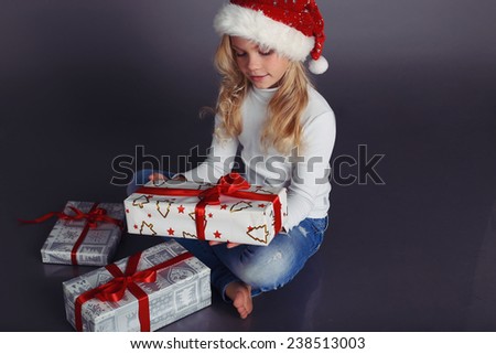 holiday photo with beautiful little girl in Santa hat and jeans and a white sweater smiles and holding a box with Christmas presents and sitting in a fashion studio