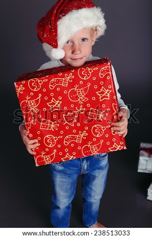 holiday photo with  little boy in Santa hat and jeans and a white sweater smiles and holding a box with Christmas presents and posing in a fashion studio