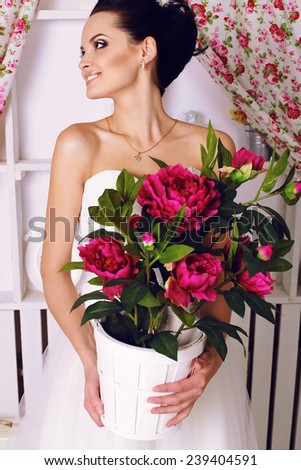 festive wedding photo of chic sexy bride in white evening dress with brunette hair posing at home near the cabinet, holding a peonies bouquet