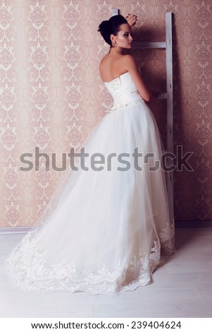 festive wedding photo of chic sexy bride with brunette hair in white evening dress with lace corset posing in the studio with ladder