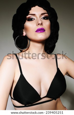 fashion photo of sexy brunette woman in wig, black lingerie and earring with bright makeup and purple lips posing in studio