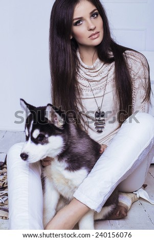 Studio photo of beautiful sexy brunette women with long straight hair wearing in cream sweater, white trousers, necklace and holding a cute little puppy of Husky
