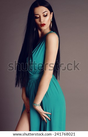 fashion photo of beautiful woman with sexy body, dark long hair wearing in green - blue evening dress with a slit,earrings,bracelet posing at studio, the wind blowing her hair