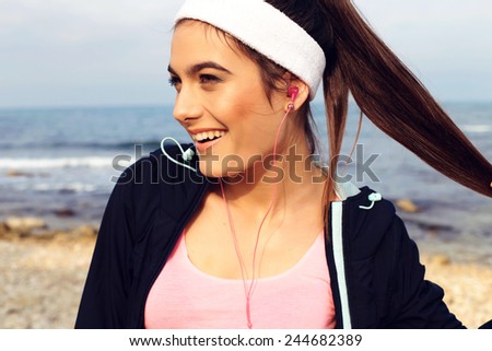 sport outdoor photo of beautiful young brunette woman in black sport suit with headband smiling and listening to music on headphones by the sea