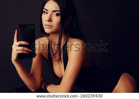 Fashion portrait of sexy tanned Asian lady with dark long hair wearing black lingerie and beautiful gold jewelry, holding a leather clutch and posing at studio