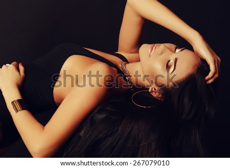Fashion portrait of sexy tanned Asian lady with dark long hair bright evening makeup, wearing black lingerie and beautiful gold jewelry lying and posing at studio