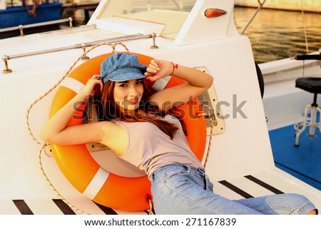 fashion outdoor photo of sexy beautiful brunette girl wearing blue jeans,top and cap smiling and relaxing on a boat with lifebuoy