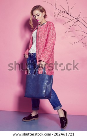 Fashion photo of sexy beautiful woman with blond curly hair, bright makeup wearing a red coat,blue jeans, holding a bag and posing on a pink background around the trees