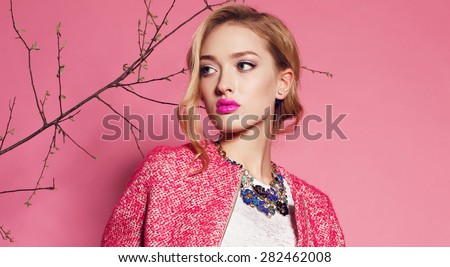 Fashion photo of sexy beautiful woman with blond curly hair, bright makeup wearing a red coat,floral necklace and posing on a pink background around the trees