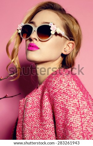 Fashion photo of sexy beautiful woman with blond curly hair, bright makeup wearing a red coat,sunglasses with floral decor and posing on a pink background