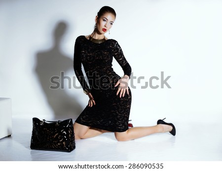 Fashion portrait of elegance brunet woman and slim sexy body wearing black lace dress with gold jewelry holding a leather bag and posing at studio
