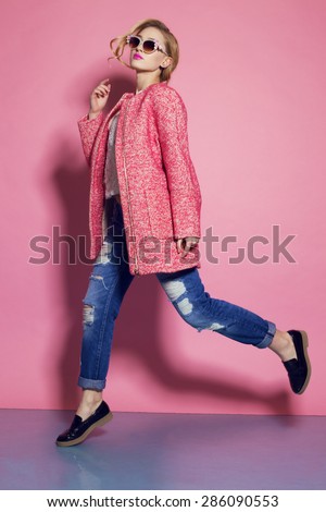 Fashion photo of sexy beautiful woman with blond curly hair, bright makeup wearing a red coat,blue jeans moving on a pink background