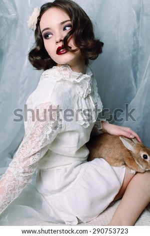Fashion photo of beautiful young woman with curly hair wearing a retro style in white lace dress, pointe shoes, holing a rabbit and posing at studio