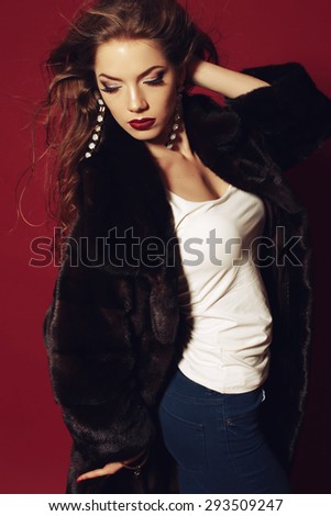 Fashion portrait of sexy lady in blue jeans and black fur coat with long blond curly hair, bright evening makeup in beautiful jewellery,  posing at studio