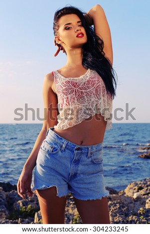 Fashion outdoor photo of sexy beautiful woman with dark hair and slim sexy body wearing a top with jeans shorts and posing at summer beach