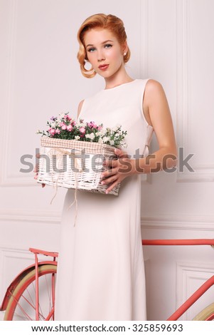 fashion photo of elegant and sexy red woman in white dress, red shoes with beautiful hairstyle posing at studio with classic interior near the bicycle and holding a box with wildflowers