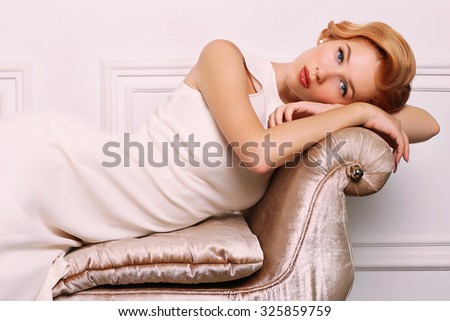 fashion photo of elegant and sexy red woman in white dress, red shoes with beautiful hairstyle lying on a luxurious couch and posing at studio with classic interior