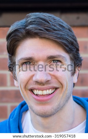 Headshot of a young man, 25-30, stood against a brick wall.  He has blue eyes, and brown hair, styled in a parting.  He is smiling.