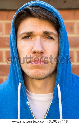 Headshot of a young man, 25-30, stood against a brick wall. He has blue eyes, and brown hair styled in a parting.  He has on a blue hoodie, and has the hood over his head.