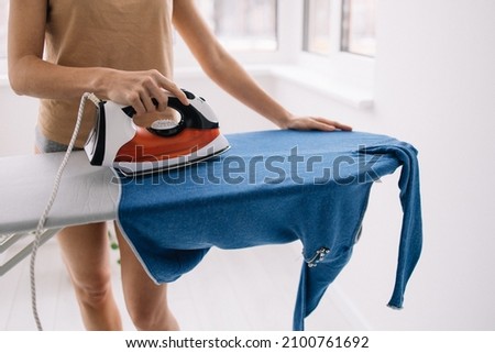 Female hands ironing blue clothes with iron on ironing board. Detail of hands holding the iron, ironing washed, wrinkled clothes