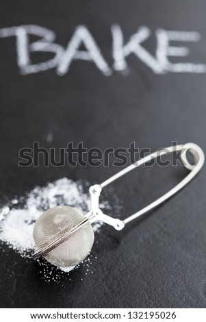 Closeup of sieve and icing sugar with word bake written in chalk on dark textured background