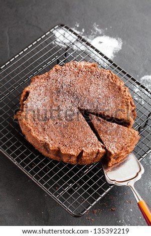 Freshly baked homemade chocolate cake on metal cooling rack with cut slice and cake server utensil on dark background