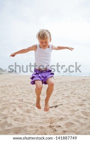 Happy barefoot young girl in purple skirt and white top jumping in sand on beach