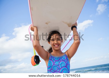 Cute smiling young girl holding pink surfboard on head at the beach with ocean and sky in background