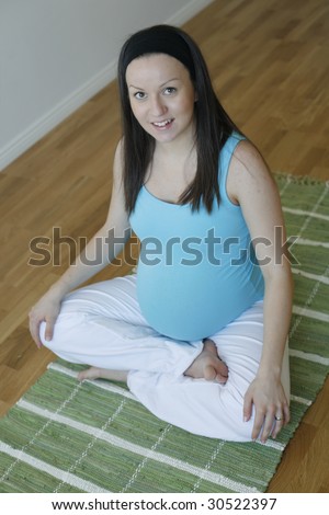 A young pregnant woman wearing a blue top and white pants in a cross-legged sitting meditative and relaxing pose on a green mat and looking at the camera.