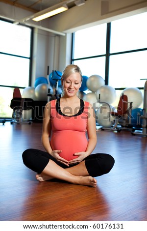 A pregnant woman smiling and relaxing in a gym while holding her belly.