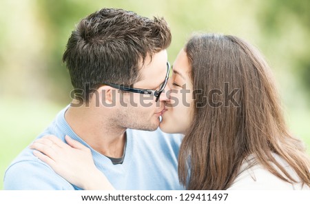Handsome man in glasses and pretty woman kissing outdoors. Husband and wife giving kiss to each other in park. Happy family expressing feelings. Portrait of beautiful romantic couple in love.