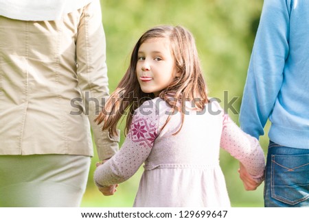 Family walking in the park. Cute child holding hands of parents. Pretty girl turning back and showing tongue. Joyful daughter playing with mother and father outdoor. Funny facial expression of kid.