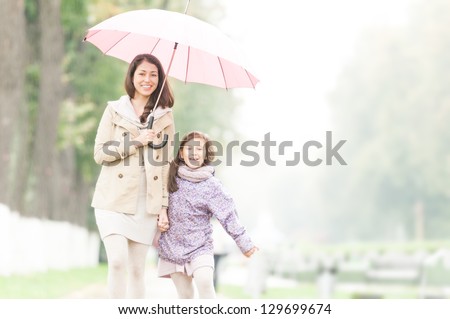 Young beautiful woman with pretty little daughter walking together in park under umbrella. Mother and daughter holding hands. Friendly family being happy and cheerful. Family walk outdoor in rain.