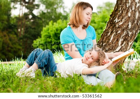 young mother sitting in grass under tree and reading book to her small daughter who is lying on knees of her mother and smiling (focus on child)