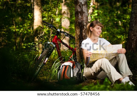 beautiful young woman sitting near her bicycle under the tree in forest with map in her hands