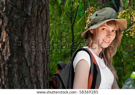 beautiful young woman standing near the tree in forest with backpack and bottle of water, smiling and looking in camera