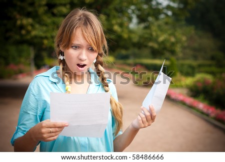 beautiful young student girl shocked by message she is reading. She is standing in campus park, holding paper with message and envelope.