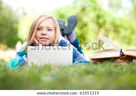 beautiful young student girl lying on grass with laptop and books, looking into the camera and smiling