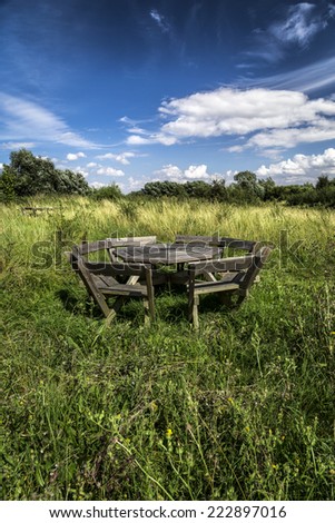 Blue sky with white fluffy clouds and a weathered wooden picnic table in uncultivated meadow in summer