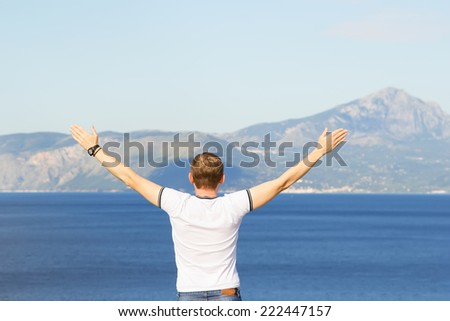 Man seen from behind, looking seaward or ocean. The man raised his hands up against the mountains. Male tourist traveler raised his hands up. Joy of freedom, the fresh sea air. Everything is fine.