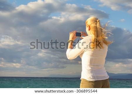 Woman photographed with your mobile phone. She was standing with his back to the camera on the background of the sea or ocean. Woman photographs the beautiful scenery of sea sunset. Be trips, vacation