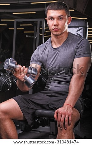Young strong muscular man sitting on a bench holding a dumbbell. A young bodybuilder in the gym. A young man with a gorgeous body in training. Processing in the style of the magazine Eksvayr.