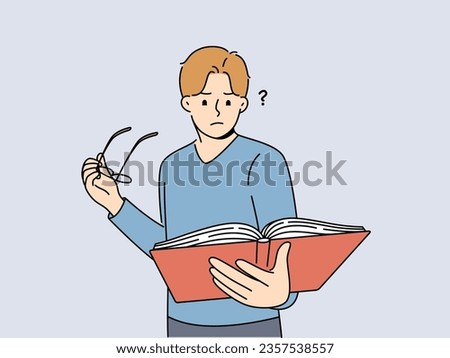 Man with book takes off glasses, trying to make out text in textbook and getting upset because of deterioration of vision. Guy is experiencing vision problems associated with excessive eye strain.