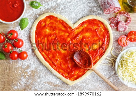 Heart shaped pizza dough covered with pizza sauce on wooden table  with cherry tomatoes,tomato sauce,mozzarella cheeses,salami,olive oil and basil leaves.Cooking with love concept for Valentine's day
